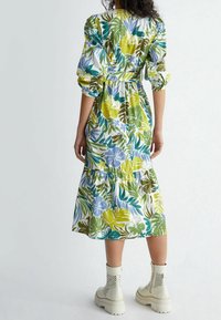 Vestido midi floral con mangas abullonadas, que presenta patrones en verde, azul y amarillo. Con volantes en el dobladillo de la falda. Combinado con botas tobilleras blancas.