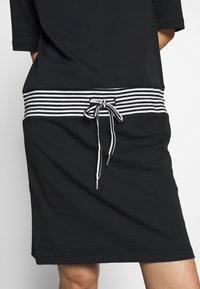 Vestido negro con una cinturilla de tela a rayas. Presenta un cordón con puntas de metal. Mangas cortas y un ajuste relajado. Textura suave.