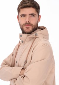 Beige wasserdichte Jacke mit Kapuze, ausgestattet mit kontrastierenden Kordelzügen, Druckknöpfen und einer glatten, strukturierten Oberfläche.