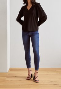 Kvinna iklädd en svart långärmad blus, blå skinny jeans och svarta stroppiga högklackade sandaler som står på ett trägolv.