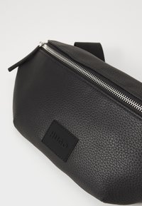 Black leather bag with a textured surface, silver zipper detail, and a rectangular logo patch. Compact design, featuring a curved shape.