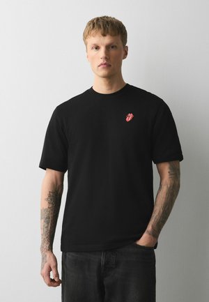 Ung mand med kort hår iført en sort t-shirt med et lille rødt tunge-logo, stående med den ene hånd i lommen mod en lysegrå baggrund.