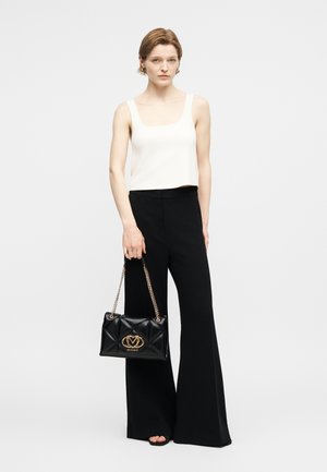 Γυναίκα με κοντά μαλλιά που φοράει λευκό αμάνικο τοπ και μαύρο παντελόνι με φαρδιά μπατζάκια, κρατώντας μαύρη τσάντα χειρός με χρυσή αλυσίδα.