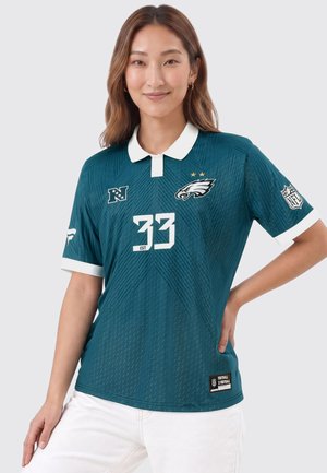 Femme portant un maillot de football des Philadelphia Eagles bleu sarcelle avec le numéro 33, col blanc, logo NFL sur la manche, et un pantalon blanc, debout avec la main sur la hanche.