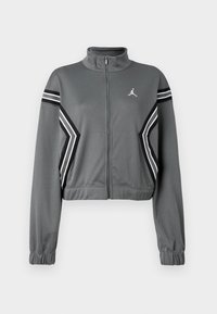 Veste de survêtement grise, légèrement cintrée, en tissu lisse avec un col montant, fermeture zippée et rayures noires et blanches sur les manches. Logo sur la poitrine.