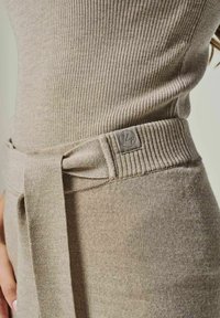 Rippstrick-beiges Kleid mit einem Gürtel in der Taille und subtiler Logo-Patch. Der Stoff ist weich, hat eine glatte Textur und eine taillierte Passform.