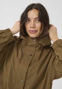 Bruine waterdichte jas met een hoge kraag, elastische manchetten en drukknoopsluiting. Glad textuur met een relaxte, oversized pasvorm en capuchon.