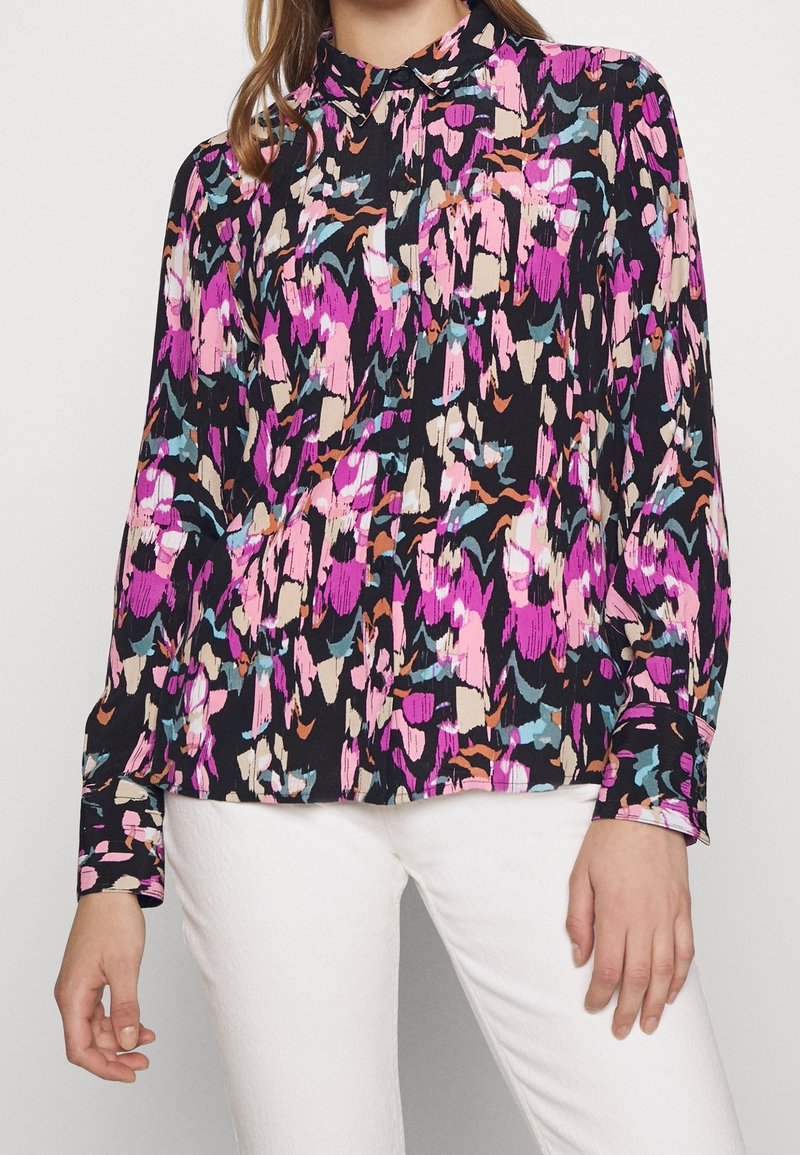 Femme portant un chemisier à manches longues boutonné avec un motif abstrait coloré rose, noir et bleu, associé à un pantalon blanc.