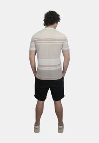 Camicia beige a maniche corte con righe orizzontali, indossata con pantaloni corti neri. Textura semplice, vestibilità casual, vista da dietro.