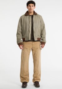 Giacca bomber verde con un collare marrone texturizzato, abbinata a pantaloni cargo beige e stivali da lavoro marroni. L'outfit presenta cuciture dettagliate.