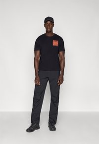 Zwarte T-shirt met korte mouwen met een oranje abstracte zakontwerp, gecombineerd met donkergrijze cargobroeken en zwarte wandelschoenen.