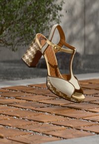 Μπεζ δερμάτινα σανδάλια με χοντρό τακούνι, σχέδιο τύπου T-πέδιλο, γεωμετρικό μοτίβο στο τακούνι και χρυσά αγκράφες. Ορατή υφή στη σόλα.