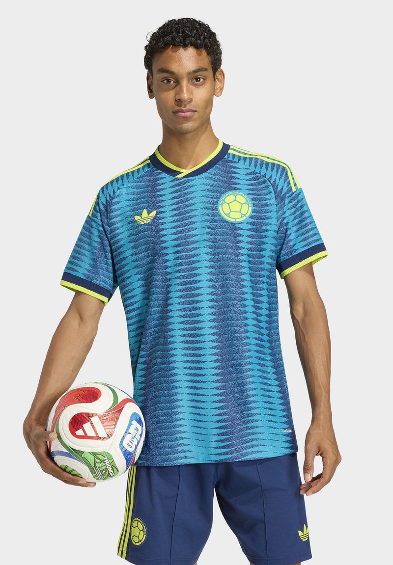 Giovane uomo con maglia da calcio blu a fantasia e pantaloncini che tiene con una mano un pallone da calcio Adidas multicolore.