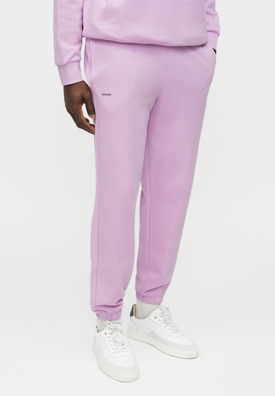Osoba ubrana w jasnofioletowe spodnie dresowe z elastycznymi mankietami i białe tenisówki, stojąca z jedną ręką w kieszeni.