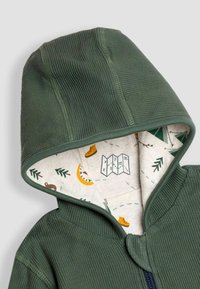 Zaļš kapuces jakas ar teksturētu ārējo slāni. Iekšpuse ir no viegla auduma ar krāsainiem kempinga tematiskajiem rakstiem. Ar rāvējslēdzēju.