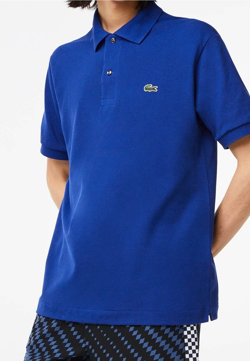 Personne portant un polo bleu à manches courtes avec un petit logo crocodile vert et un pantalon noir et bleu à motifs.