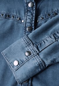 Camisa de mezclilla azul clara con botones a presión, que presenta una tela texturada y costuras visibles. Los puños son ligeramente más anchos con bordes redondeados.