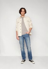 Jasnobeżowa koszula-żakiet na szarym polo, zestawiona z niebieskimi dżinsami i czarnymi trampkami. Prosty design z zapięciami na guziki i kieszeniami.