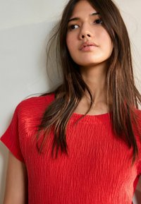 Haut rouge texturé avec un col rond et des manches courtes, présentant un motif de tissu froissé. Cheveux longs et raides, encadrant le visage.