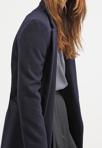 Mörkblå kappa i mjukt tyg, med en elegant design som har öppen framsida, något figursydd form och långa ärmar.