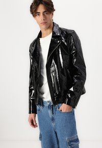 Sort patentlæder motorcykeljakke med lynlåslommer og et elegant design, båret over en hvid skjorte og parret med blå jeans.