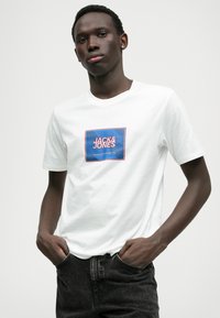 Biela bavlnená tričko s modrým a červeným obdĺžnikovým grafickým motívom s textom "JACK&JONES". Krátke rukávy a klasický strih okrúhleho výstrihu.