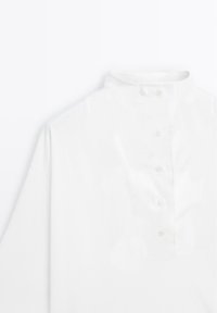 Chemise blanche à col montant avec cinq petits boutons alignés verticalement sur le devant près du col, vue de l'angle du buste supérieur.