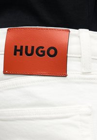 Κοντινό πλάνο λευκών τζιν από τζιν με ένα κόκκινο δερμάτινο έμπλαστρο ραμμένο στη μέση, όπου αναγράφεται το όνομα της μάρκας «HUGO» με μαύρα γράμματα.