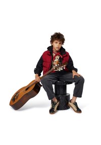 Gilet matelassé rouge et noir sur un t-shirt graphique, pantalon en denim gris et baskets noir et beige. Assis sur un tabouret noir à pois avec une guitare.