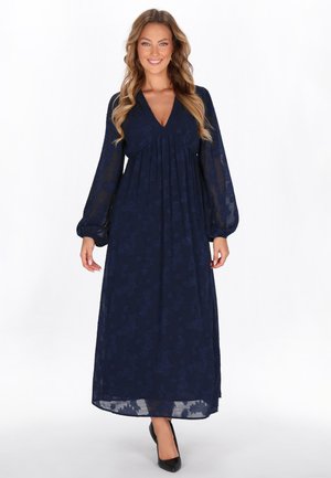 Langes marineblaues Kleid mit V-Ausschnitt, strukturiertem Blumenmuster und Puffärmeln; aus leichtem Stoff gefertigt. Getragen mit schwarzen Absätzen.