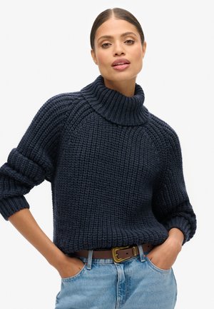 Suéter de punto azul marino con un cuello alto de canalé y mangas enrolladas. Llevado con jeans de mezclilla azul claro y un cinturón marrón, enfatizando la textura y el ajuste.