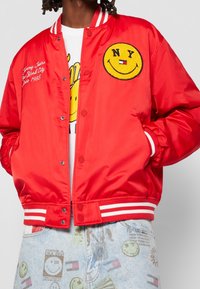 Giacca bomber in raso rosso con polsini e colletto a strisce bianche. Presenta un logo con faccina sorridente gialla e testo ricamato sul davanti.