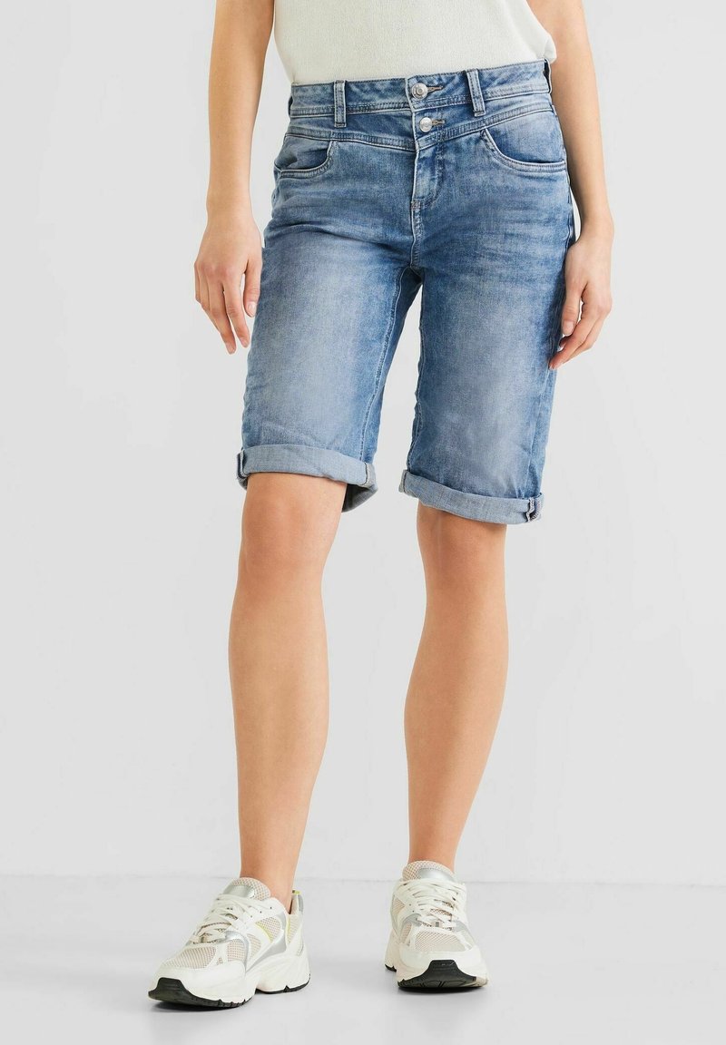Light blue denim shorts with rolled cuffs, featuring a classic five-pocket design and button fly, paired with white athletic shoes.