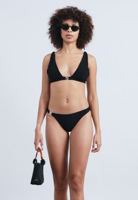 Μαύρο ριγέ μπικίνι με τριγωνικό τοπ, που διαθέτει μεταλλικό κούμπωμα, συνδυασμένο με ψηλός καβαλέτος. Το μοντέλο κρατά μια μικρή μαύρη τσάντα.