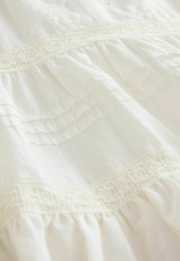Tissu en coton blanc avec des volants en couches et une broderie texturée. Présente un accent de dentelle décorative le long de la couture horizontale.