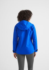 Woman with curly hair wearing a bright blue hooded jacket and gray leggings, standing facing away against a plain background.