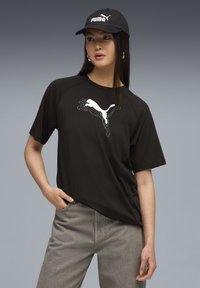 Sort t-shirt med korte ærmer med et hvidt grafisk Puma-logo på forsiden, parret med grå jeans. Stoffet virker glat og let.
