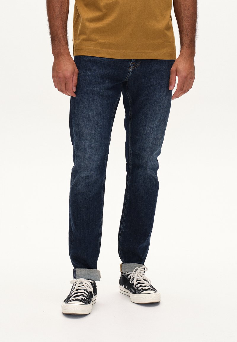 Jeans de mezclilla azul oscuro con corte recto, remangados en el tobillo, combinados con zapatillas blancas y negras. La parte superior es de color mostaza.