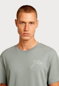 Lichtgrijs katoenen T-shirt met een ronde hals, voorzien van een subtiel logo in cursief op de linkerborst. Soepele textuur en een ontspannen pasvorm.