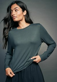 Mørkegrøn langærmet top med et struktureret stof, rund halsudskæring og løs pasform, parret med en sort nederdel. Stoffet viser en subtil ribbing.