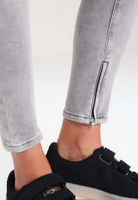 Pantalones de mezclilla gris ajustados, con una cremallera en el tobillo. Combinados con zapatos negros con correas de gancho y detalles perforados.