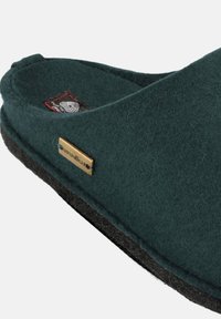 Green felt slipper with a black sole, featuring a gold metal logo accent, open design, and soft texture.