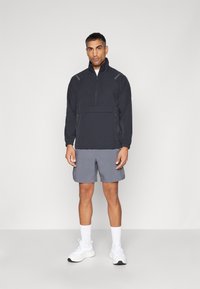 Zwarte ritsjack met een hoge kraag, gecombineerd met grijze shorts en witte sneakers. De jas heeft zijritsen en reflecterende accenten.