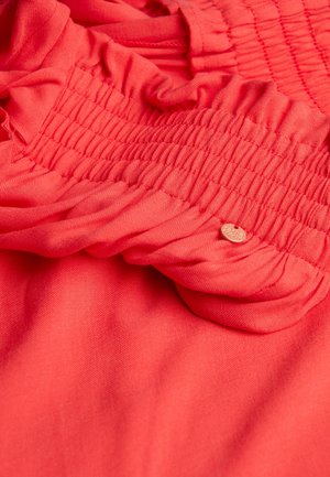 Coral fabric with gathered texture and ruffle details; features a small, tan heart-shaped tag. Soft, smooth material.