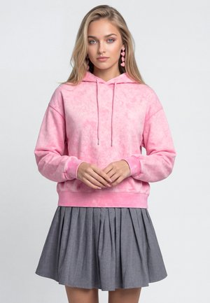 Ung kvinde med langt blond hår iført pink tie-dye hættetrøje, hjerteformede øreringe og en plisseret grå nederdel, der står foran en ensartet baggrund.