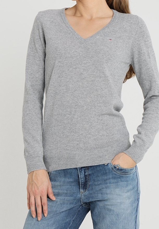 Femme portant un pull gris clair à col en V et manches longues et un jean bleu avec une main dans la poche, sur un fond uni.