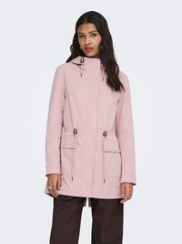 Femme aux longs cheveux bruns portant une veste à capuche rose clair avec cordons et grandes poches à rabat, associée à un pantalon brun foncé.