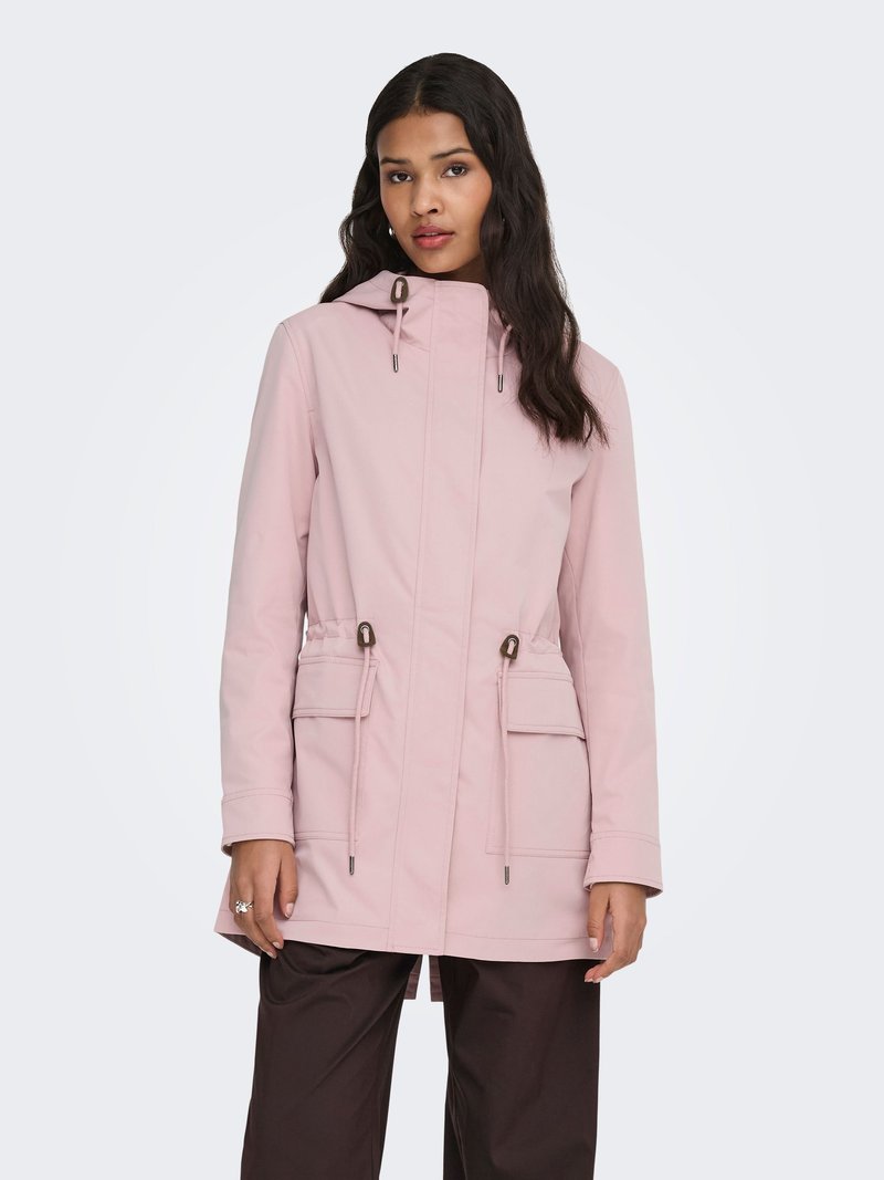 Femme aux longs cheveux bruns portant une veste à capuche rose clair avec cordons et grandes poches à rabat, associée à un pantalon brun foncé.