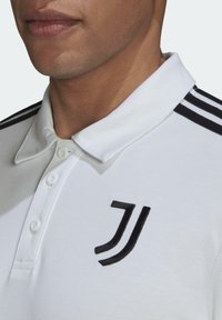 Gros plan d'une personne portant un polo blanc avec des bandes noires sur l'épaule et un logo "JJ" noir sur la poitrine.