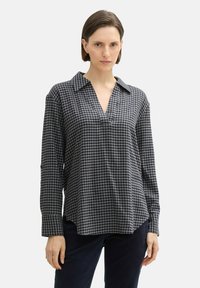 Femme portant une chemise à manches longues, ample, à carreaux noirs et gris avec un col en V, associée à un pantalon sombre.